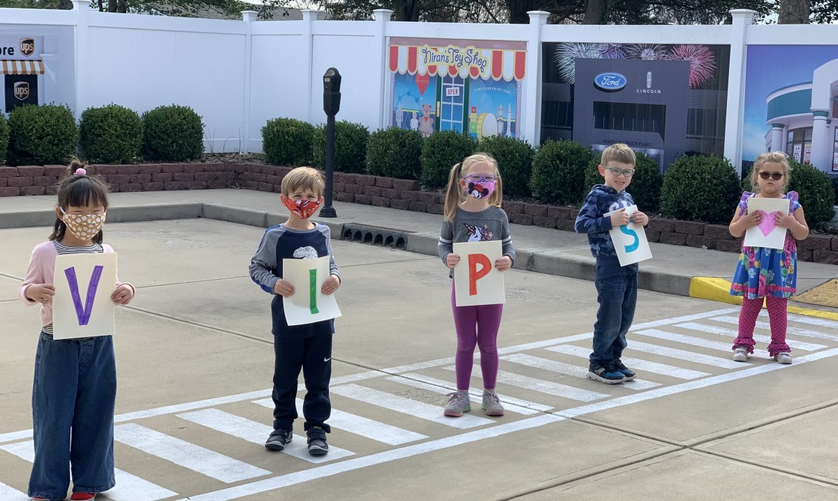 Ms. Ashley's class stand outside on the crosswalk at Kids Town Preschool, holding papers that spell out V-I-P-S and a pink heart. From left to right, Millie, Nate, Emma, Gabe and Lane.